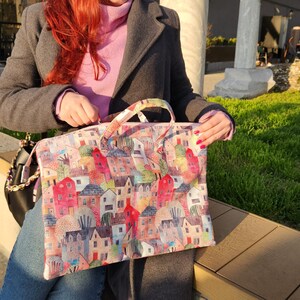 Op de afbeelding: Een roze en witte tote bag met een kleurrijke illustratie van huizen en bomen. De tas heeft twee bovenste handvatten en een ritssluiting.