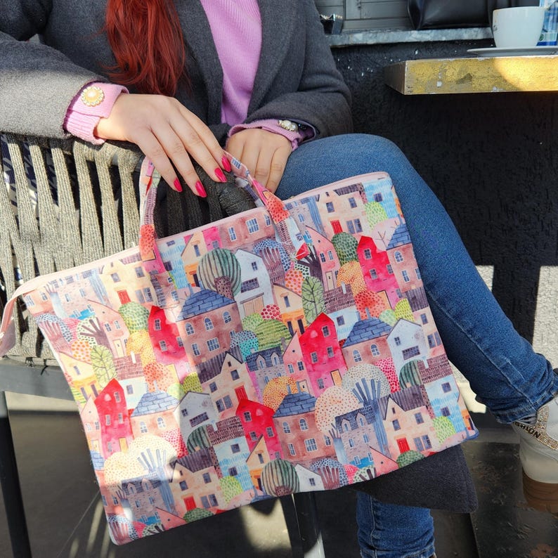 Op de afbeelding: Een roze tote bag met een kleurrijke illustratie van een stad met huizen, bomen en andere details. De tas heeft twee handvatten en een ritssluiting.