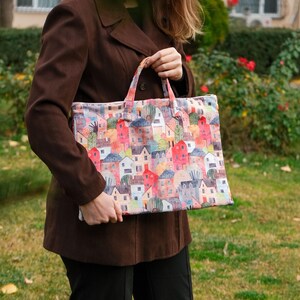 Op de afbeelding: Een bruine tote bag met een kleurrijke print van huizen en bomen. De tas heeft twee handvatten en is gemaakt van stevige stof.