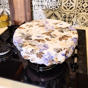 Op de afbeelding: Een witte en paarse bloemenstofhoes voor een pot of pan op een zwart fornuis.