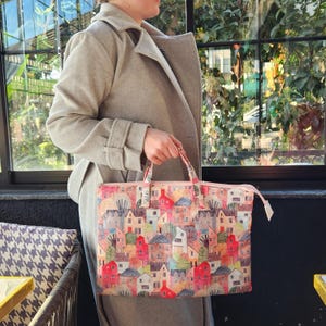 Op de afbeelding: Een roze en witte tote bag met een kleurrijke print van huizen en bomen. De tas heeft een ritssluiting en een bovenste handvat.