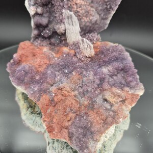 May include: A close-up of a natural mineral specimen featuring a cluster of purple, red, and green crystals. The crystals have a textured surface and are arranged in a layered formation. A light-colored crystal formation is in the center.