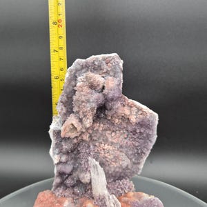 May include: A natural amethyst geode specimen with a ruler showing a height of approximately 8 inches. The geode features a cluster of small, sparkling purple crystals and a base of reddish-brown material. The background is a dark gray.