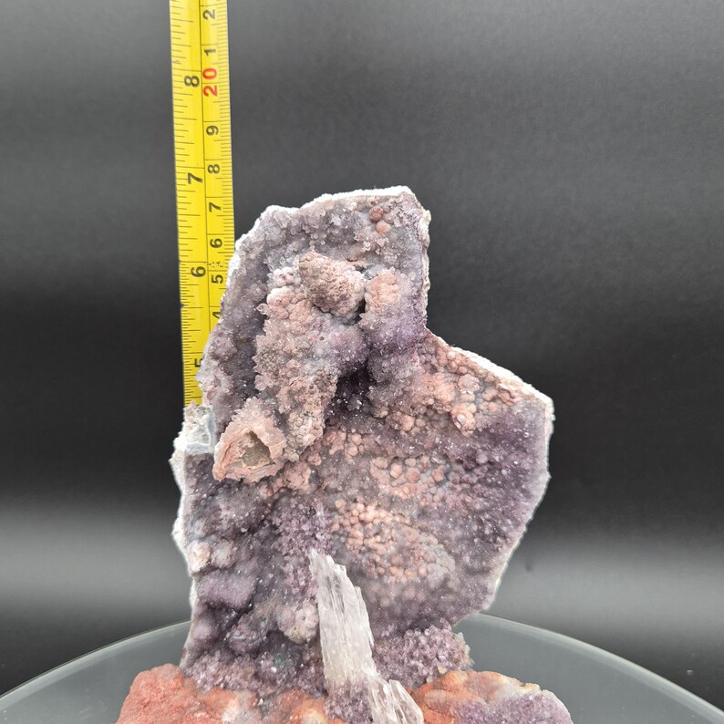 May include: A natural amethyst geode specimen with a ruler showing a height of approximately 8 inches. The geode features a cluster of small, sparkling purple crystals and a base of reddish-brown material. The background is a dark gray.