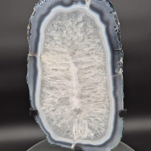 May include: A polished agate slice, showcasing a natural pattern of concentric bands in shades of gray, white, and translucent crystal. The stone is displayed on a black stand, revealing its intricate internal structure and unique geological formation.