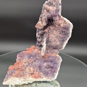 May include: A natural amethyst geode specimen with a cluster of purple crystals, accented by reddish-brown and white mineral formations. The geode is displayed on a reflective surface, showcasing its unique texture and color variations.