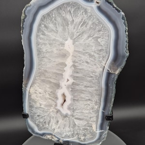 May include: A polished agate slice with a natural, organic shape. The stone features bands of gray, white, and blue hues, with a central crystalline structure. The agate is displayed on a stand, showcasing its unique patterns and textures.