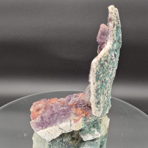 May include: A natural mineral specimen featuring a cluster of purple, pink, and green crystals. The crystals are set against a white and green textured matrix, creating a striking contrast. The specimen is displayed on a reflective surface.