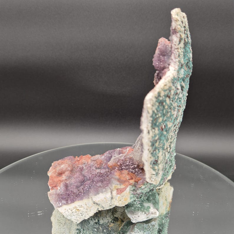 May include: A natural mineral specimen featuring a cluster of purple, pink, and green crystals. The crystals are set against a white and green textured matrix, creating a striking contrast. The specimen is displayed on a reflective surface.