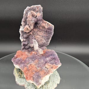 May include: A natural amethyst crystal cluster with a mix of purple, pink, and orange hues. The crystal formation has a rough, textured surface and a small, white crystal protruding from the center. The base is a mix of green and gray.