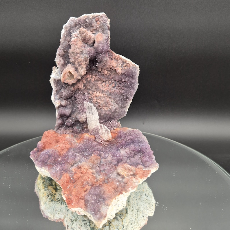 May include: A natural amethyst crystal cluster with a mix of purple, pink, and orange hues. The crystal formation has a rough, textured surface and a small, white crystal protruding from the center. The base is a mix of green and gray.