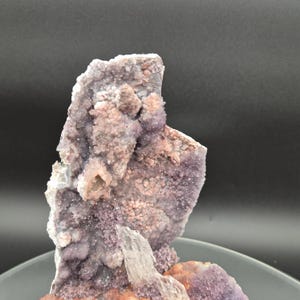 May include: A close-up of a natural amethyst geode with a crystalline structure. The geode features a mix of purple, pink, and white hues, with a rough, textured surface. The crystal formation is visible, showcasing the natural beauty of the mineral.