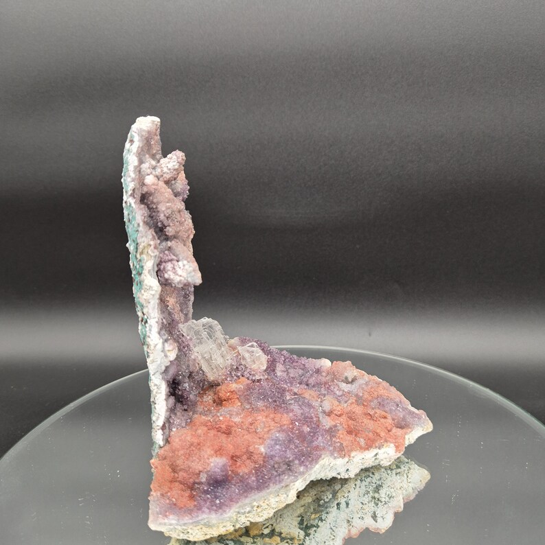 May include: A natural mineral specimen featuring a cluster of purple, pink, and white crystals. The crystals have a rough, textured surface and are arranged in a layered formation. The specimen is displayed on a reflective surface.
