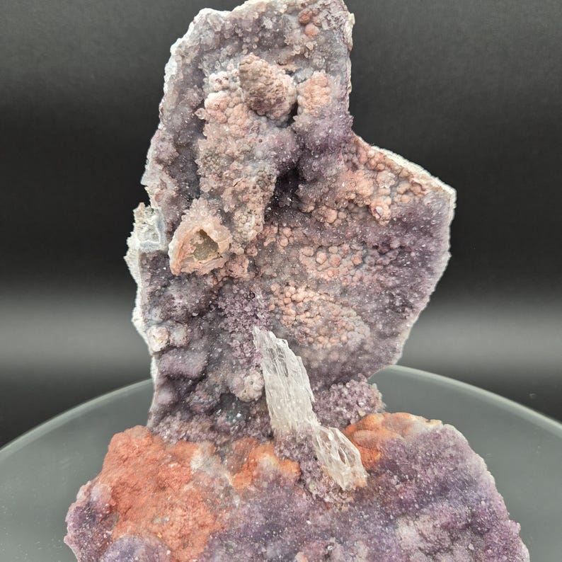 May include: A natural amethyst geode specimen with a crystalline structure. The geode features a mix of purple, pink, and reddish-brown hues. A clear, elongated crystal formation is visible, adding contrast to the textured surface. The geode is displayed on a gray surface.