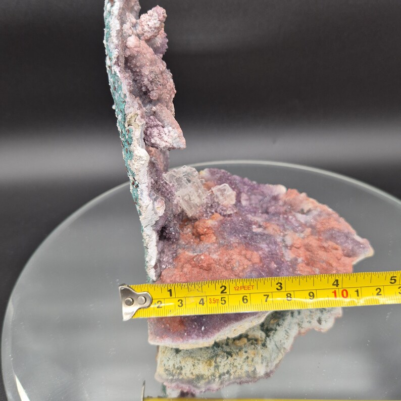 May include: A natural mineral specimen featuring a cluster of purple, pink, and white crystals. The specimen is approximately 4 inches wide, with a tape measure providing scale. The mineral has a rough, textured surface and a mix of colors.