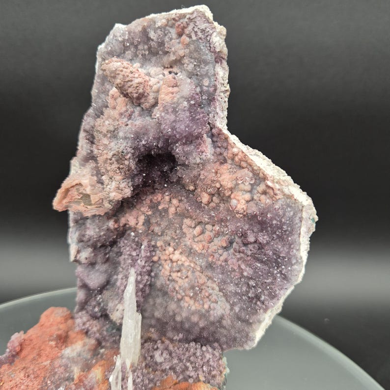 May include: A close-up of a natural mineral specimen. The rock formation features a mix of colors, including shades of purple, pink, and white. The surface has a textured, crystalline appearance, with a prominent crystal formation in the center.