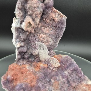 May include: A natural mineral specimen featuring a cluster of purple, pink, and reddish-brown crystals. A clear, elongated crystal is embedded in the center. The specimen has a rough, textured surface and is displayed on a clear, circular base.