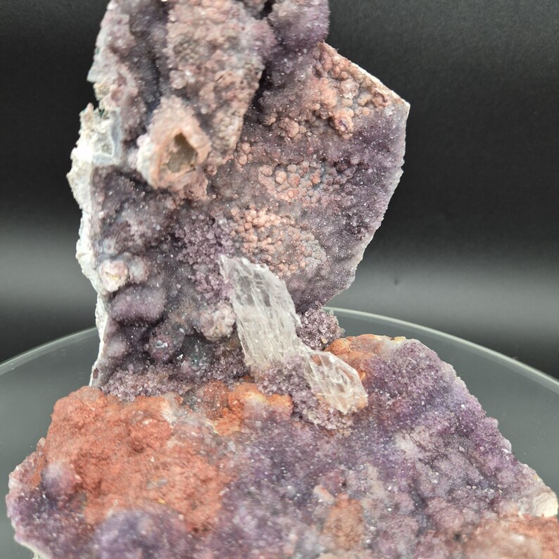 May include: A natural mineral specimen featuring a cluster of purple, pink, and reddish-brown crystals. A clear, elongated crystal is embedded in the center. The specimen has a rough, textured surface and is displayed on a clear, circular base.