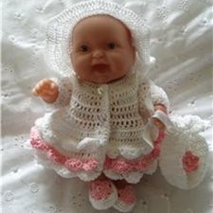 May include: A white crocheted baby doll outfit with a pink trim. The outfit includes a dress, cardigan, hat, shoes, and a small bag.