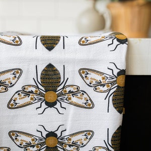 May include: White kitchen towel with a repeating pattern of black and gold bees with floral details.