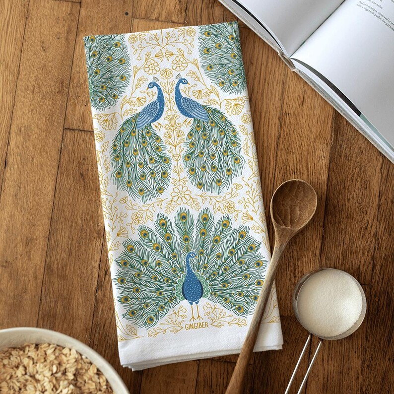 May include: A white kitchen towel with a blue and green peacock design. The towel features a repeating pattern of peacocks with their tails spread out. The text "GINGBER" is printed in the bottom right corner of the towel.
