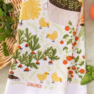 May include: White kitchen towel with a colorful garden-themed design. The towel features illustrations of carrots, tomatoes, yellow chicks, and the word "GINGIBER". The design is set against a white background with green and orange accents.