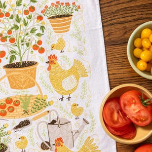 May include: White kitchen towel with a garden-themed design. The illustration features tomato plants, chickens, and gardening tools in shades of orange, yellow, and green. Two bowls of tomatoes are on a wooden surface.