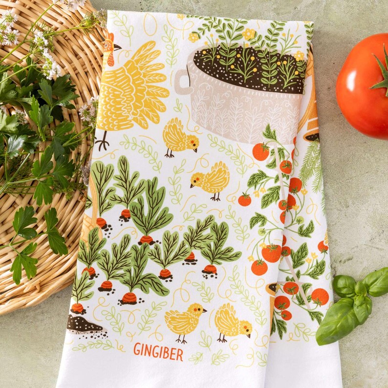 May include: White kitchen towel with a colorful garden-themed design. The towel features illustrations of carrots, tomatoes, chickens, and a flower pot. The word "GINGIBER" is printed at the bottom. The towel is folded and placed next to a basket of herbs and a tomato.