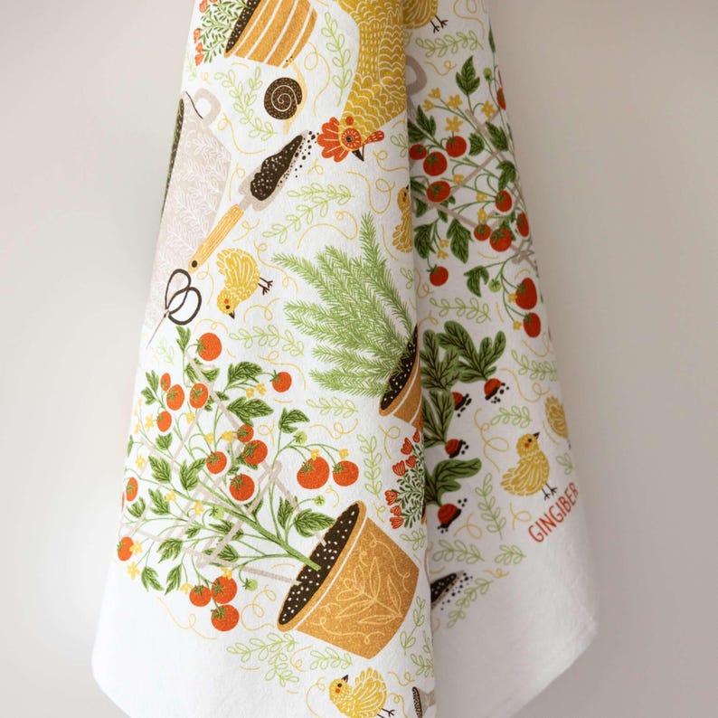 May include: White kitchen towel with a colorful garden-themed print. The design includes potted tomato plants, gardening tools, yellow chicks, and the word "Gingiber". The towel is made of a soft, absorbent fabric.