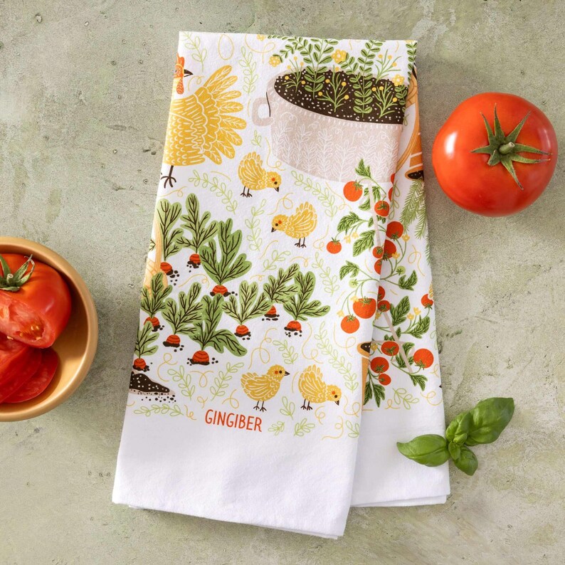 May include: White kitchen towel with a colorful garden-themed design. Features illustrations of carrots, tomatoes, chickens, and a watering can. The word "GINGIBER" is printed on the bottom. A fresh tomato and basil sprig are also visible.