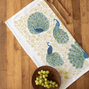 May include: White kitchen towel featuring a peacock design. The peacock has blue and green feathers and is surrounded by yellow floral accents.