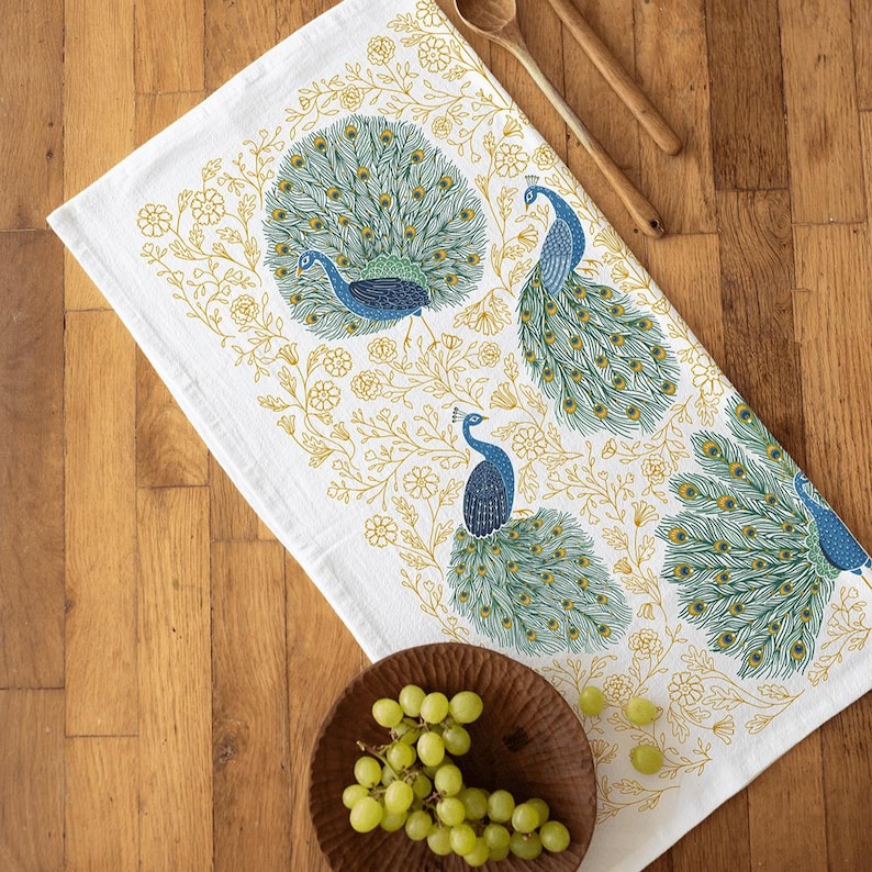 May include: White kitchen towel featuring a peacock design. The peacock has blue and green feathers and is surrounded by yellow floral accents.