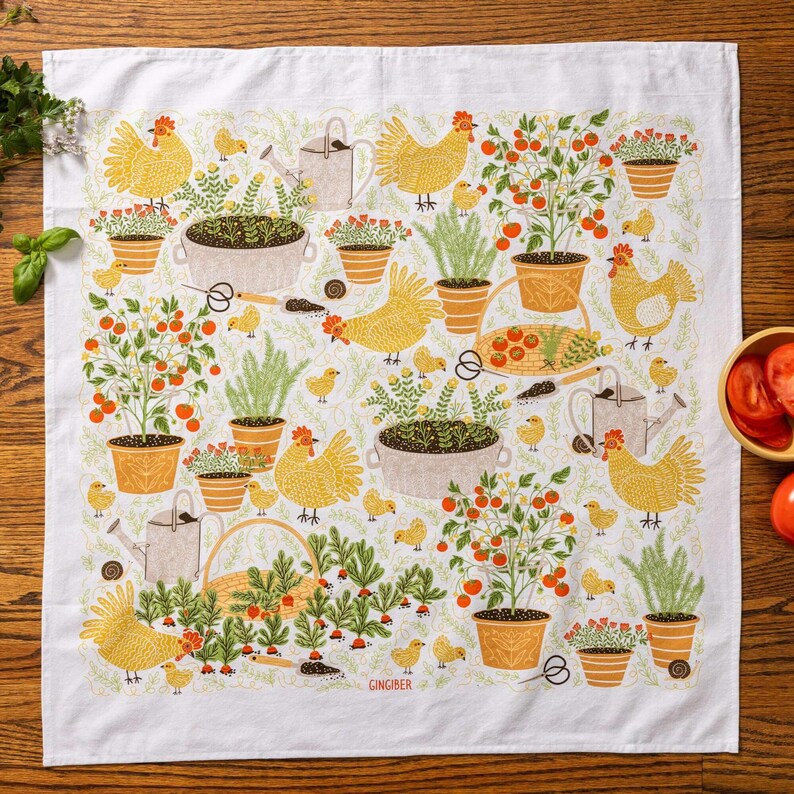 May include: White kitchen towel with a garden-themed design. The pattern includes yellow chickens, tomato plants, potted flowers, watering cans, and gardening tools. The towel has a border of green foliage and the word "GINGIBER" is printed on the bottom.