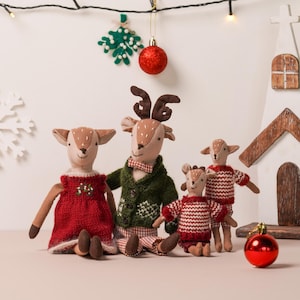Op de afbeelding: Een gezin van vier pluche herten die feestelijke kersttruien en jurken dragen. De herten zitten voor een witte houten kerk met een rode kerstbal.