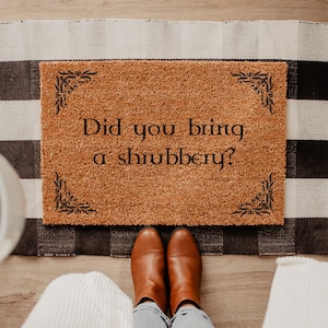 May include: A rectangular tan coir doormat featuring black decorative corner flourishes and the black text "Did you bring a shrubbery?" printed in the center. The mat is positioned on a black and white buffalo check fabric rug.