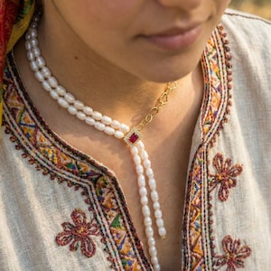 May include: A pearl necklace with a gold chain and a red gemstone square pendant. The necklace has two strands of white pearls that cascade down the chest. The gold chain has a heart-shaped design.