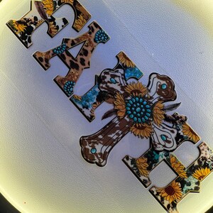 May include: Wooden cut-out letters spelling "FAITH" with a cross-shaped "H". The letters and cross feature a mix of patterns: cowhide, leopard print, sunflowers, and turquoise accents. The design has a rustic, country aesthetic.