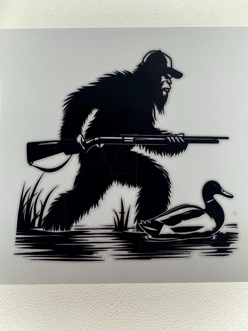 May include: Black and white graphic of Bigfoot, wearing a cap and holding a rifle, stalking a duck in water. The image is a silhouette, with details of the fur, rifle, and duck visible.