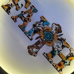 May include: Wooden sign spelling "FAITH" with a cross. The letters and cross feature a mix of patterns including sunflowers, cowhide, and leopard print. Turquoise accents are incorporated into the design, creating a rustic, country aesthetic.