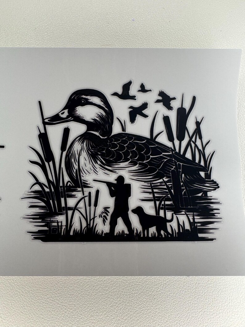 May include: Black and white silhouette of a duck in a marsh setting. The image includes a hunter aiming a rifle, a dog, flying birds, and cattails. The duck is the central focus, with detailed feather patterns.