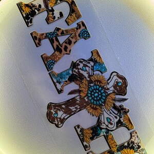 May include: Wooden cutouts spelling the word "FAITH". The letters feature a mix of patterns including cowhide, leopard print, sunflowers, and turquoise accents. The letter "H" is a cross shape with a sunflower and turquoise center.