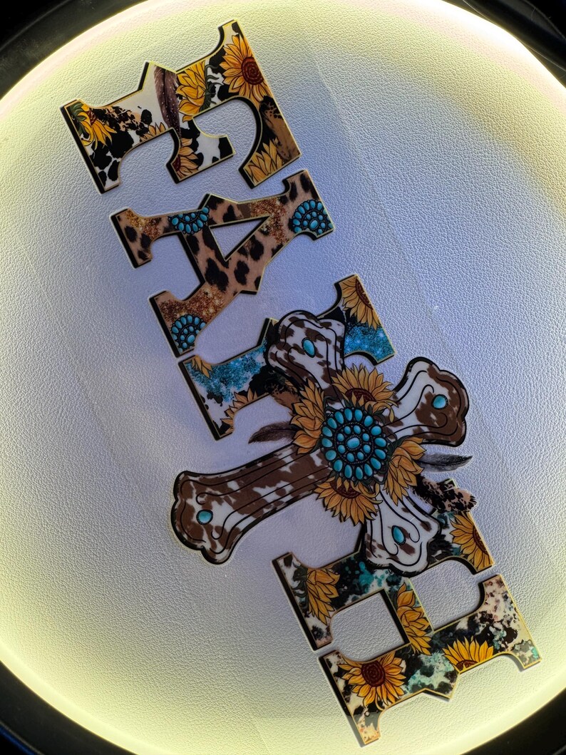 May include: Wooden cutouts spelling the word "FAITH". The letters feature a mix of patterns including cowhide, leopard print, sunflowers, and turquoise accents. The letter "H" is a cross shape with a sunflower and turquoise center.