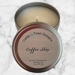 May include: A silver tin candle with a white label that reads "Cedar Town Designs Coffee Shop" and "soy wax | hand poured in Cedartown Ga 6 oz (170.1g)"