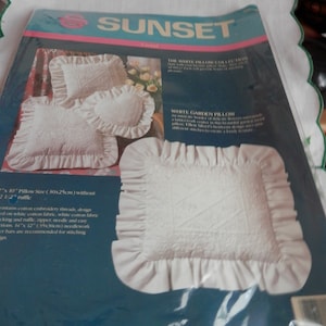 May include: A white pillow with a ruffled edge and intricate embroidery. The pillow is packaged in a plastic bag with a white and blue label that reads "Sunset" and "White Garden Pillow". The label also includes instructions for stitching the pillow.