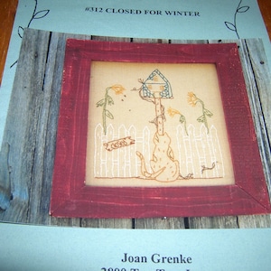 May include: A framed cross-stitch embroidery depicting a small house with a sign that reads "Closed" and a cat sitting in front of a picket fence. The embroidery is on a beige fabric with brown, blue, and white thread. The frame is a dark red wood.