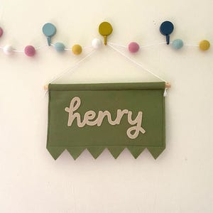 May include: A green fabric banner with the name "henry" in a light beige script. The banner has a zigzag bottom edge and is suspended from a wooden dowel. A garland of colorful felt balls hangs above.