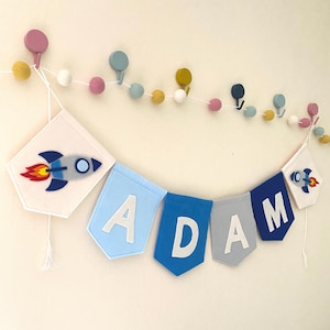 May include: A felt banner spelling "ADAM" in white letters, with a rocket ship design on each end. The banner is in shades of blue and grey, with a string of colourful felt balls above.