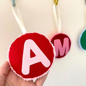 Könnte beinhalten: Roter Filzkreis mit weißem, gewelltem Rand und einem großen weißen Buchstaben "A". Ein weißes Band ist zum Aufhängen befestigt. Weitere Filzkreise mit Buchstaben sind sichtbar.