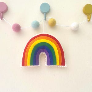 Könnte beinhalten: Bunte Filz-Regenbogen-Dekoration mit gewelltem Rand, mit roten, orangen, gelben, grünen, blauen und violetten Schichten. Eine Girlande aus pastellfarbenen Filzkugeln hängt über dem Regenbogen, befestigt an Wandhaken.