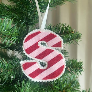 May include: A felt Christmas ornament shaped like the letter "S". The ornament features alternating red and pink stripes, a white scalloped edge, and a white ribbon for hanging. The ornament is hanging on a green Christmas tree.